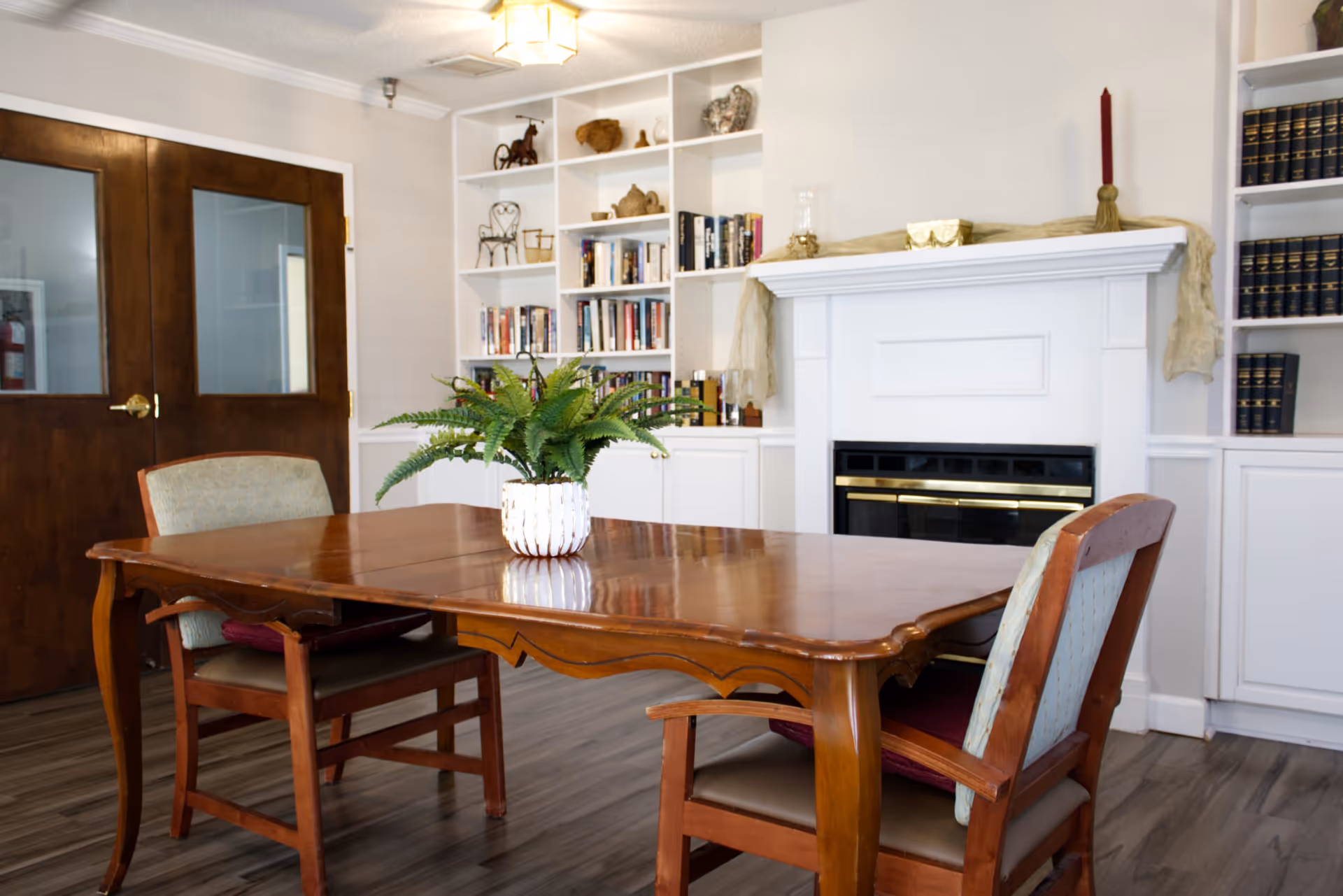 A wooden dining table with a potted plant and chairs in a communal room with a fireplace and built-in bookshelves.