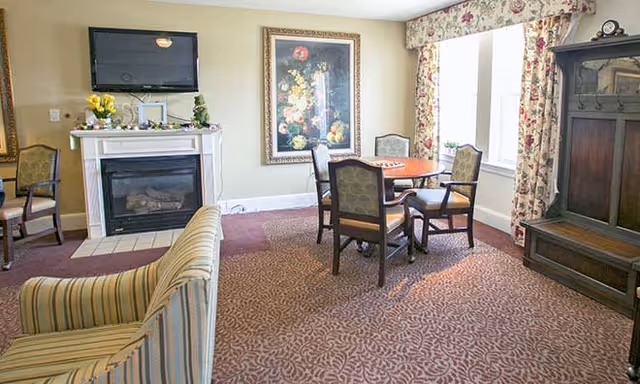 A cozy living room area with a striped sofa in the foreground, a fireplace with a TV mounted above it, a round wooden table with four chairs near a window with floral curtains, a large floral painting on the wall, and a wooden cabinet on the right side.