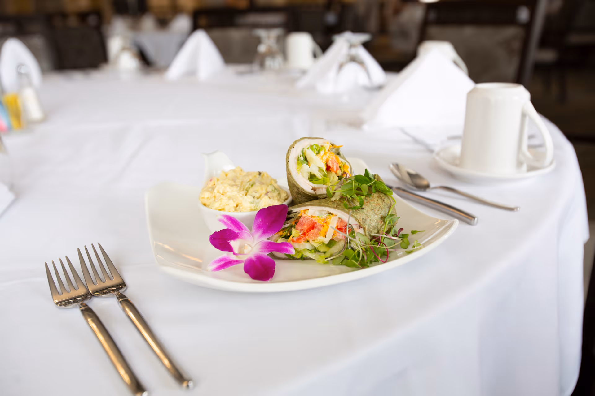 A white plate on a white tablecloth with a wrap sandwich cut in half, filled with vegetables and possibly egg, garnished with microgreens and a purple orchid flower. Next to the wrap is a small bowl of potato salad. Two forks are placed on the table to the left of the plate, and a white coffee cup with a saucer and spoon is on the right side of the plate. The background shows a dining area with tables and chairs.