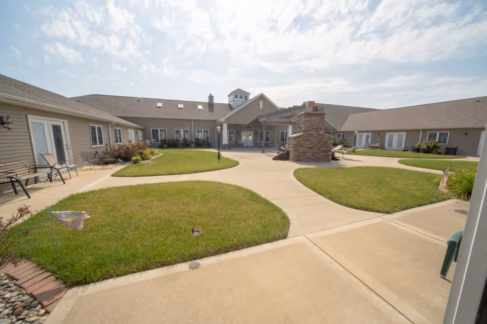 Outdoor courtyard area of a senior living facility with a central stone fireplace, surrounded by a paved walkway and green grass patches. The courtyard is enclosed by single-story buildings with beige siding and multiple windows and doors. There are benches and chairs along the building walls, and the sky is partly cloudy.