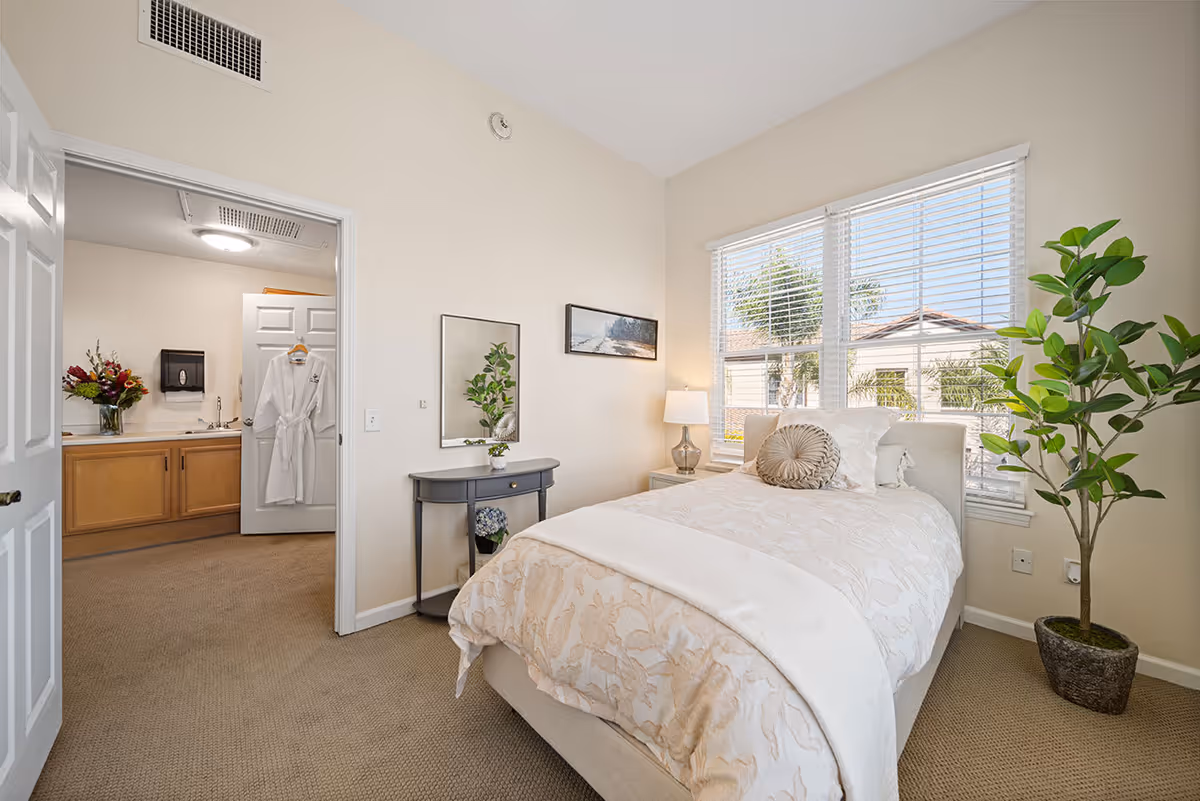 A bright bedroom with a single bed covered in a beige patterned comforter and decorative pillows. A small gray table with a plant and a mirror hangs on the wall above it. A large window with white blinds lets in natural light, showing a view of palm trees and rooftops outside. A tall potted plant is in the corner near the window. An open door leads to a bathroom area with a wooden vanity, a white bathrobe hanging on the door, and a vase of flowers on the counter.