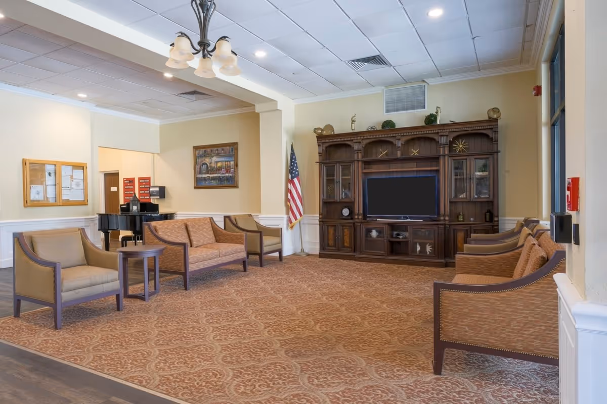 A spacious living room area in a senior living facility with patterned carpet, several cushioned chairs and sofas arranged around a large wooden entertainment center with a flat-screen TV. An American flag stands in the corner, and a black grand piano is visible in the background. The walls are light-colored with a framed painting and a bulletin board.