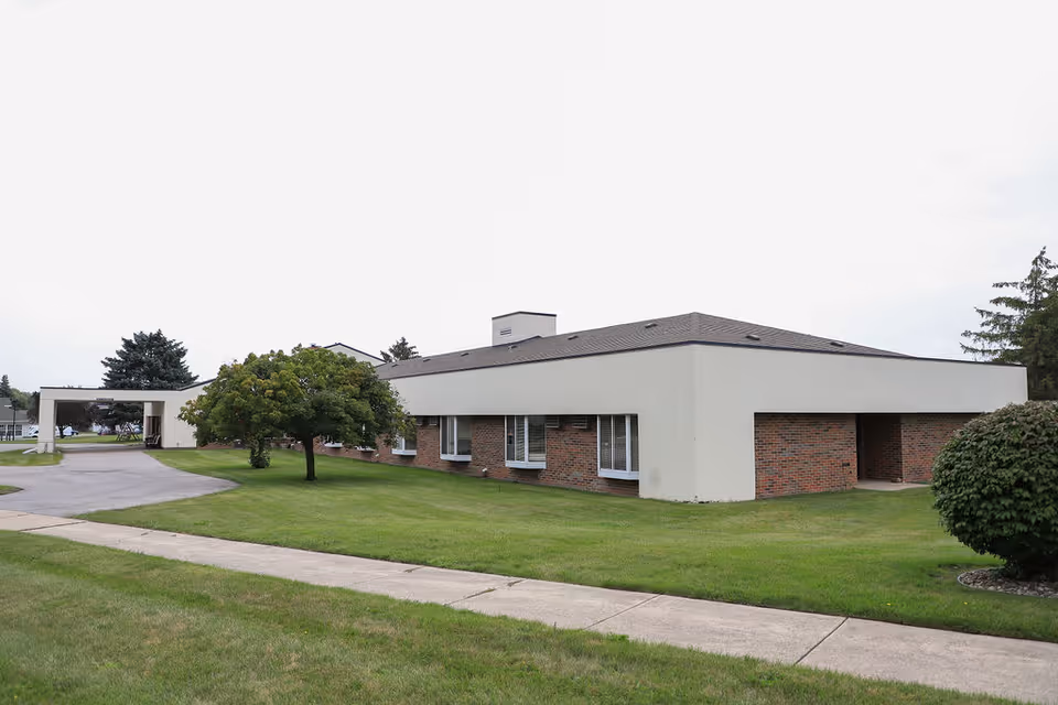 Exterior view of a single-story assisted living facility building with a combination of white and brick walls, surrounded by green grass, trees, and a paved driveway leading to a covered entrance.