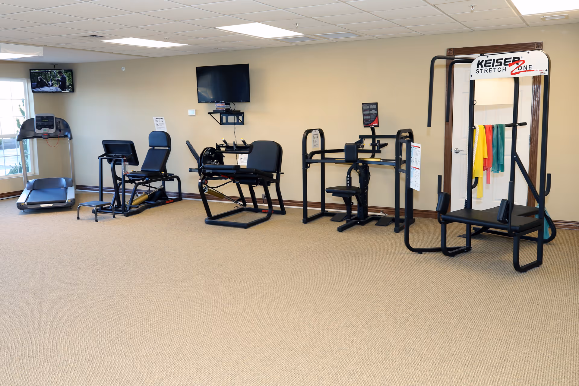 A fitness room in a senior living facility with various exercise machines including a treadmill, stationary bike, and strength training equipment. The room has beige walls, carpeted floor, a wall-mounted TV, and a window letting in natural light.