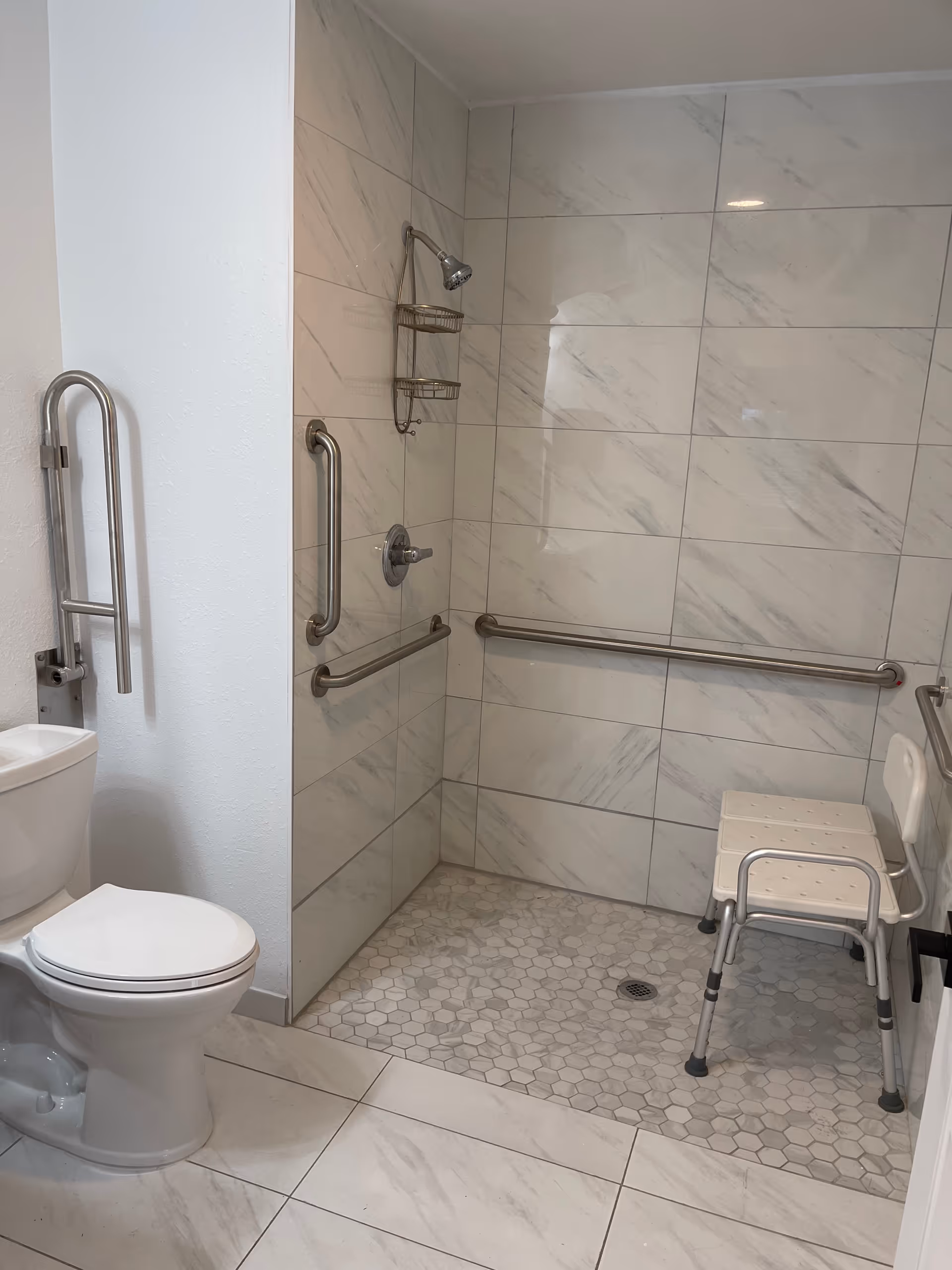 A bathroom with a white toilet and a walk-in shower area. The shower has white and gray marble-like tiles on the walls and hexagonal tiles on the floor. There are multiple stainless steel grab bars installed on the walls for support, and a white shower chair with metal legs is placed inside the shower. A small shower caddy is hanging from the showerhead.