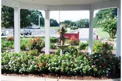 View of a garden area with a multi-tiered water fountain surrounded by blooming flowers and greenery, seen through white columns of a covered porch. In the background, there is a road with vehicles and trees.