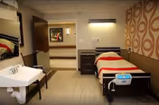 A nursing facility bedroom with a single hospital-style bed, bedside table, wall-mounted sink, and an open door leading to a hallway.