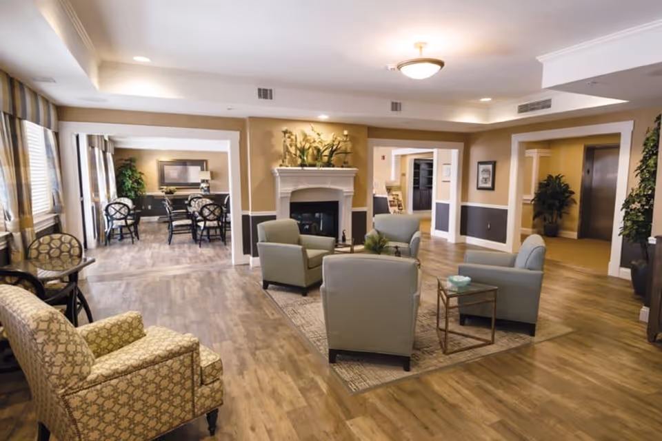 A spacious and well-lit senior living facility common area with wooden flooring, beige walls, and a white fireplace adorned with plants. The room features comfortable seating including four light green armchairs arranged around a small table on a rug, and a patterned armchair near the window. In the background, there is a dining area with a table and chairs, large windows with striped curtains, and potted plants. The area also includes an elevator and framed artwork on the walls.