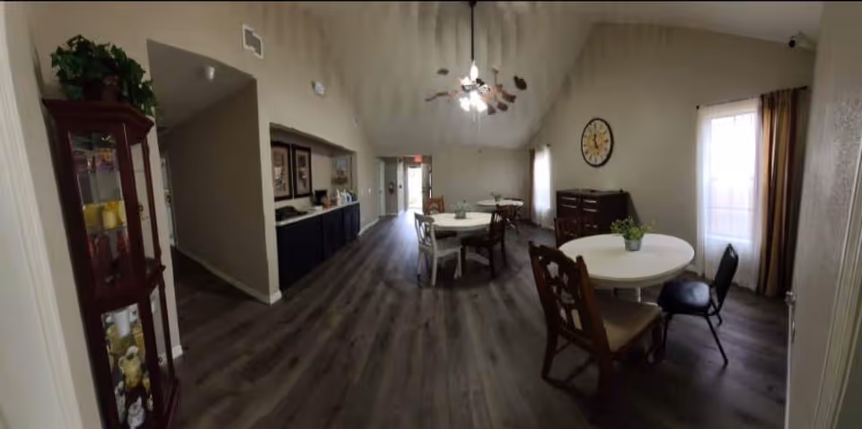 Interior view of a common area with multiple round tables and chairs arranged on a dark wood floor. There is a ceiling fan with lights, a large clock on the wall, and windows with curtains letting in natural light. A cabinet with decorative items is visible on the left side.