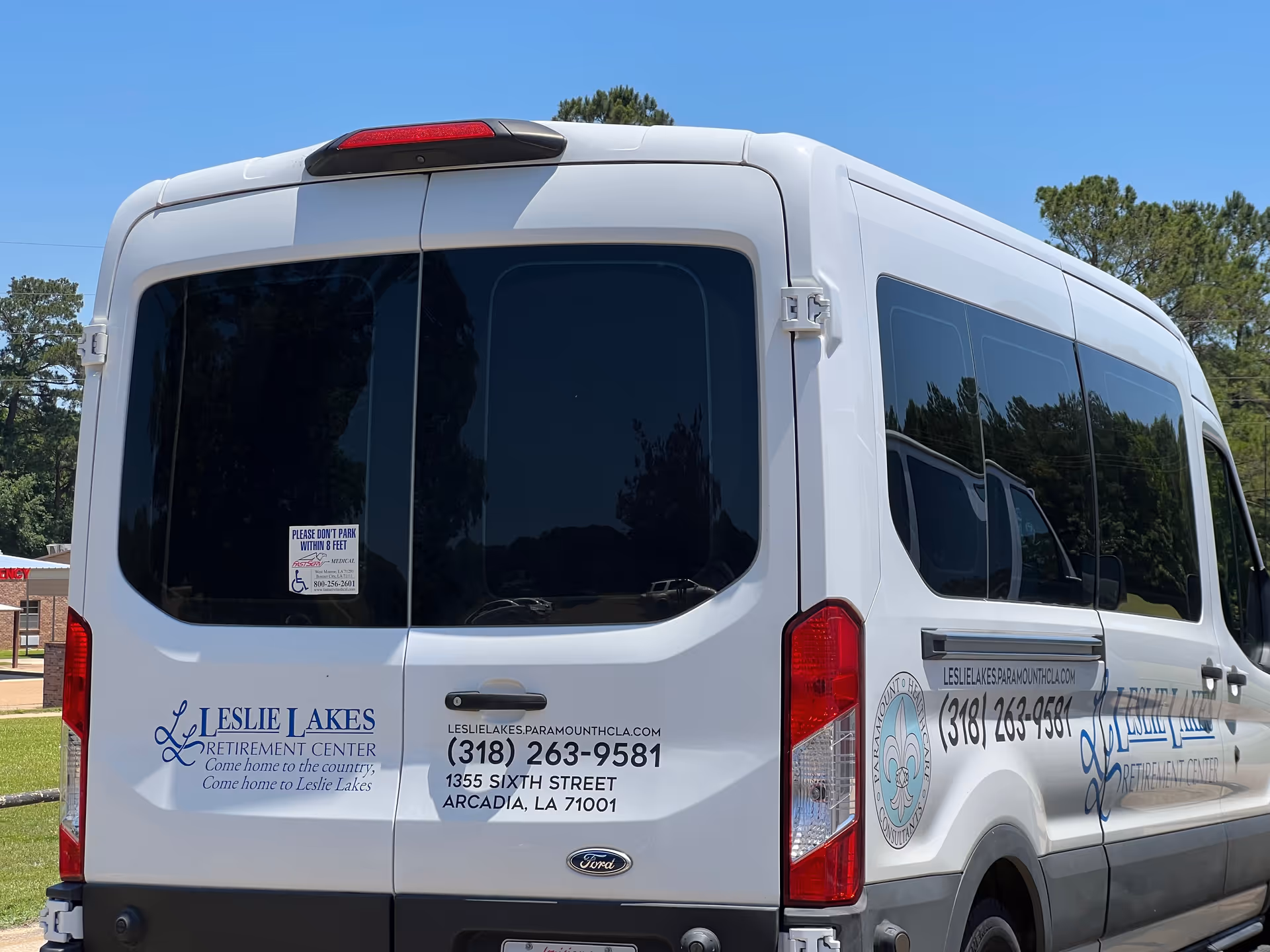 Rear and side view of a white Leslie Lakes Retirement Center shuttle van parked outdoors with signage and contact information.