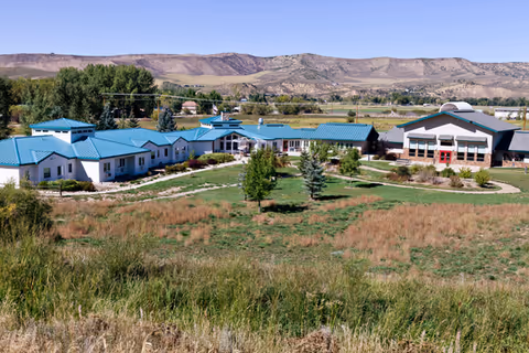 A wide exterior view of The Haven facility showing multiple single-story buildings with teal-colored roofs surrounded by green lawns and trees, set against a backdrop of rolling hills and a clear blue sky.