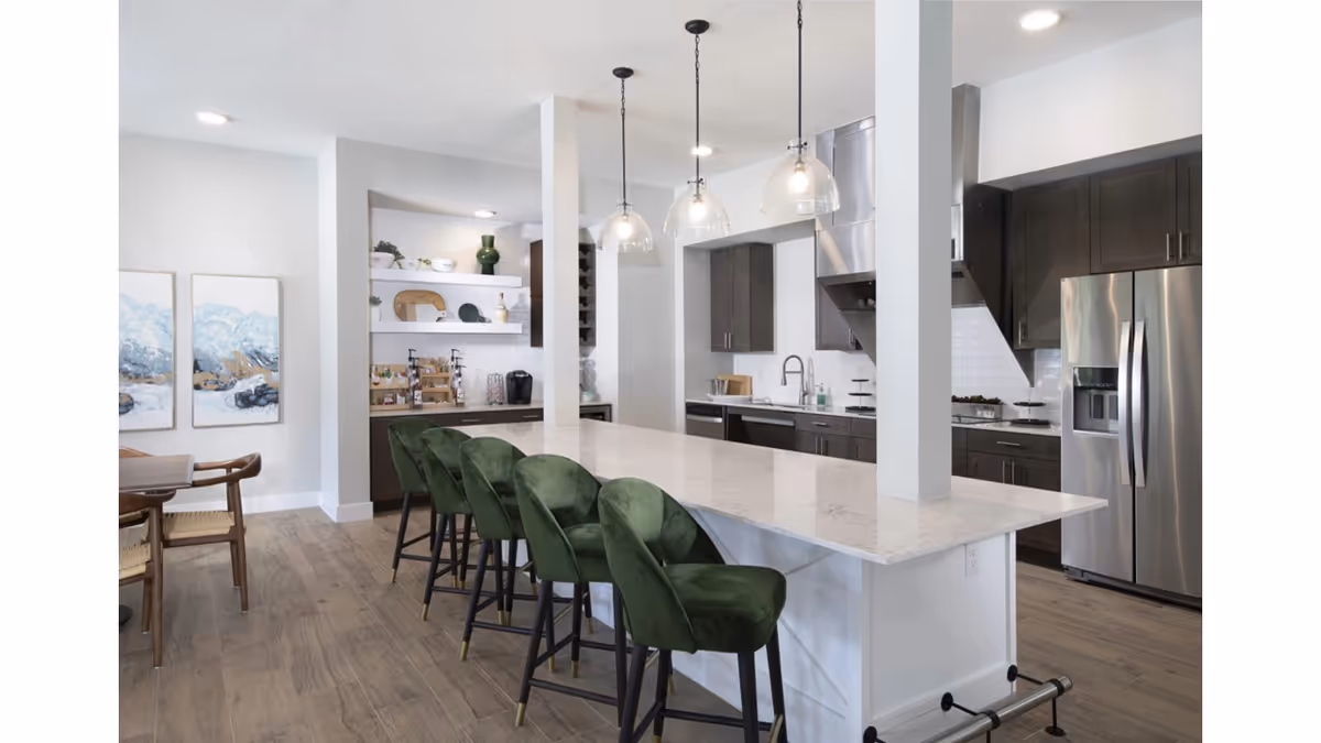 Modern kitchen interior with a long white marble island countertop surrounded by five green velvet bar stools. The kitchen features dark wood cabinets, stainless steel refrigerator, stove with a hood, and a sink. Three pendant lights hang above the island. In the background, there are shelves with decorative items and a coffee station. The floor is wooden, and two abstract paintings are visible on the left wall.