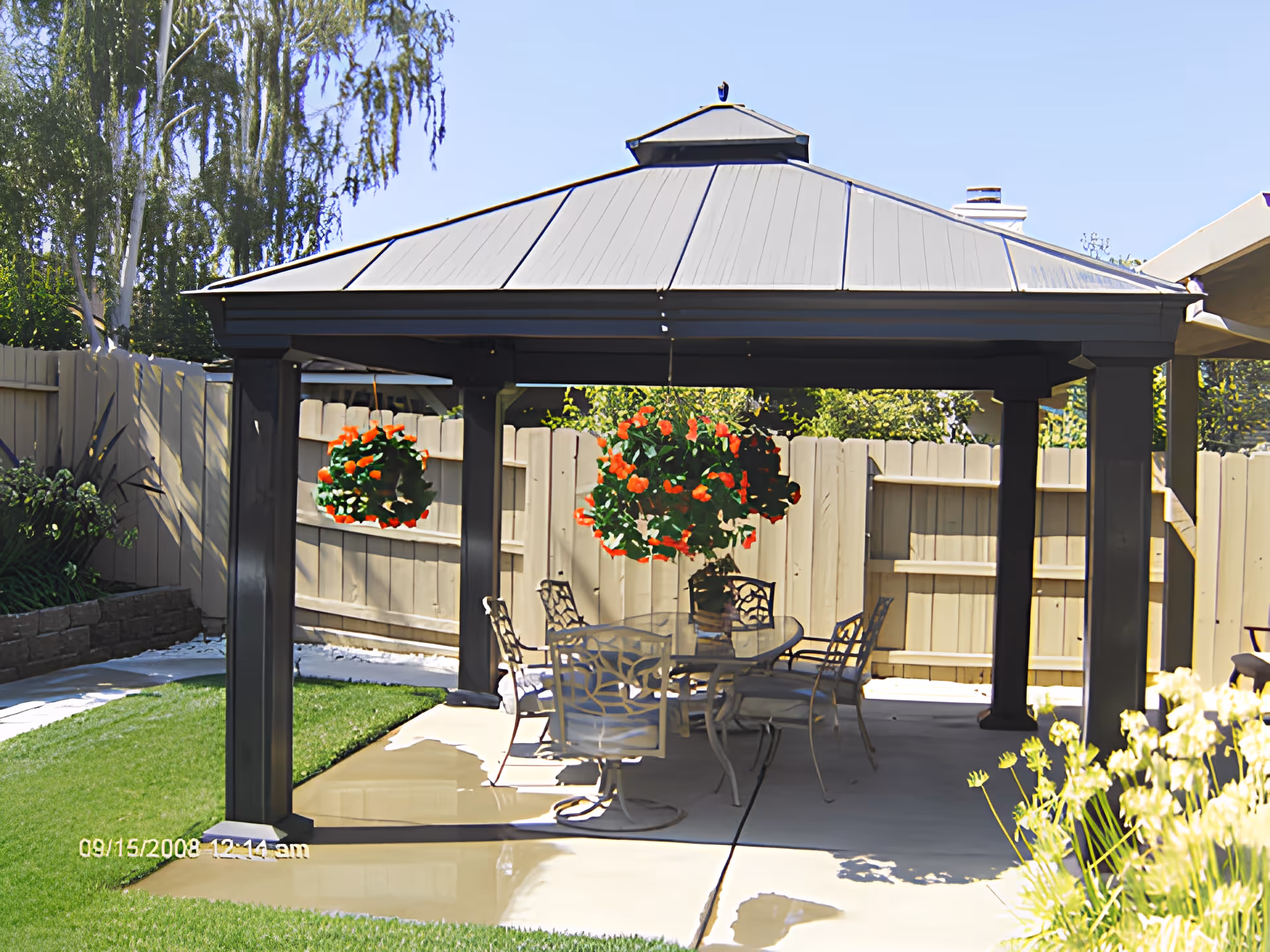 Metal gazebo in a fenced backyard with a table and chairs beneath and hanging flower baskets.