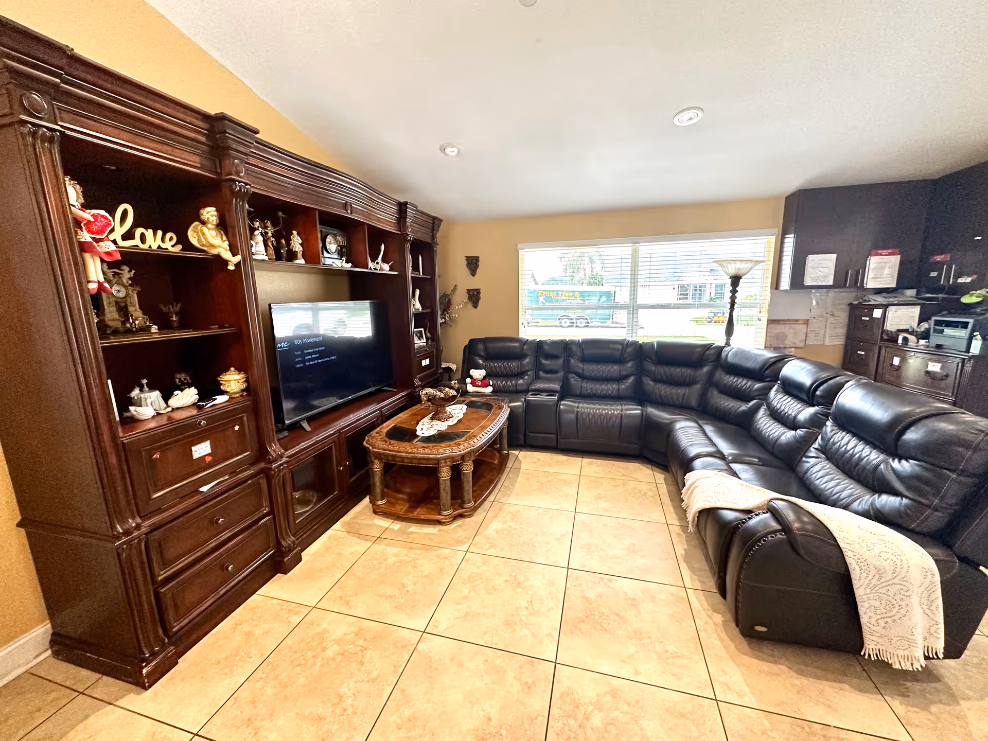 A bright living room with a large black leather sectional facing a wooden entertainment center with a TV, a coffee table, and a window behind.