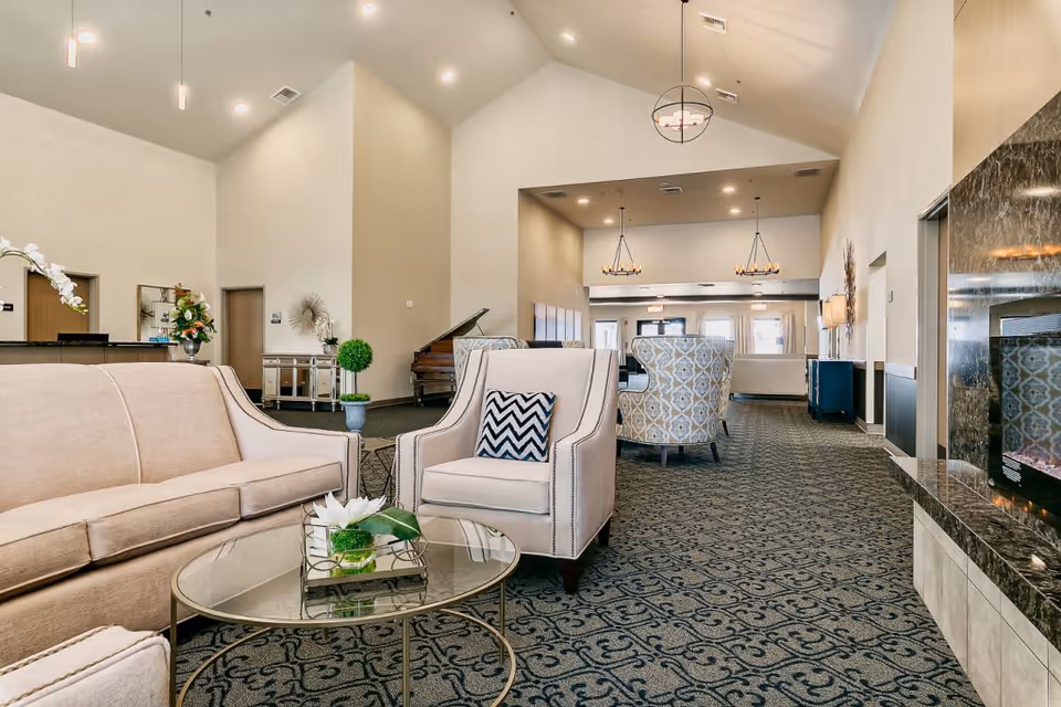 Spacious senior living lounge with sofas, armchairs, a glass coffee table, patterned carpet, and a fireplace.