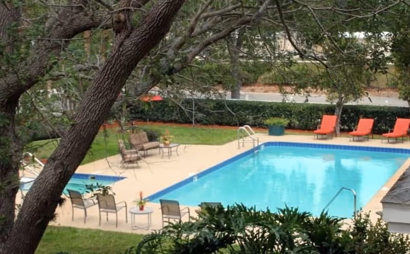 Outdoor swimming pool area with several lounge chairs and patio furniture surrounded by trees and greenery.
