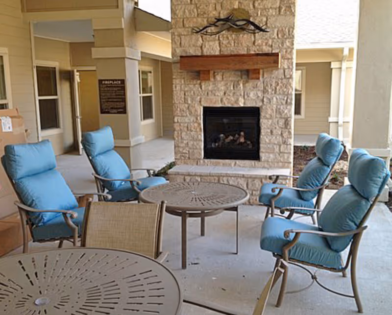 Outdoor covered patio area with a stone fireplace, four cushioned chairs with blue cushions arranged around a round metal table, and another round metal table in the foreground.