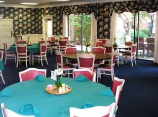 Dining room with round tables covered in teal tablecloths, pink-and-white chairs, floral wallpaper, and large windows/doors looking out to a patio.