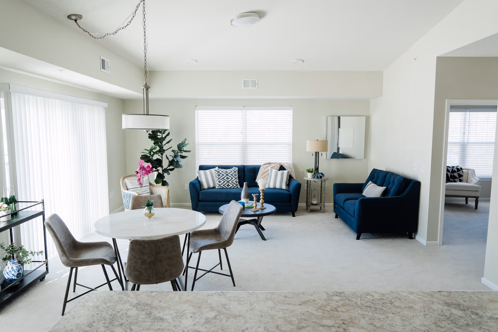 Bright open living and dining area with two blue sofas, a round white dining table, and large windows with blinds.
