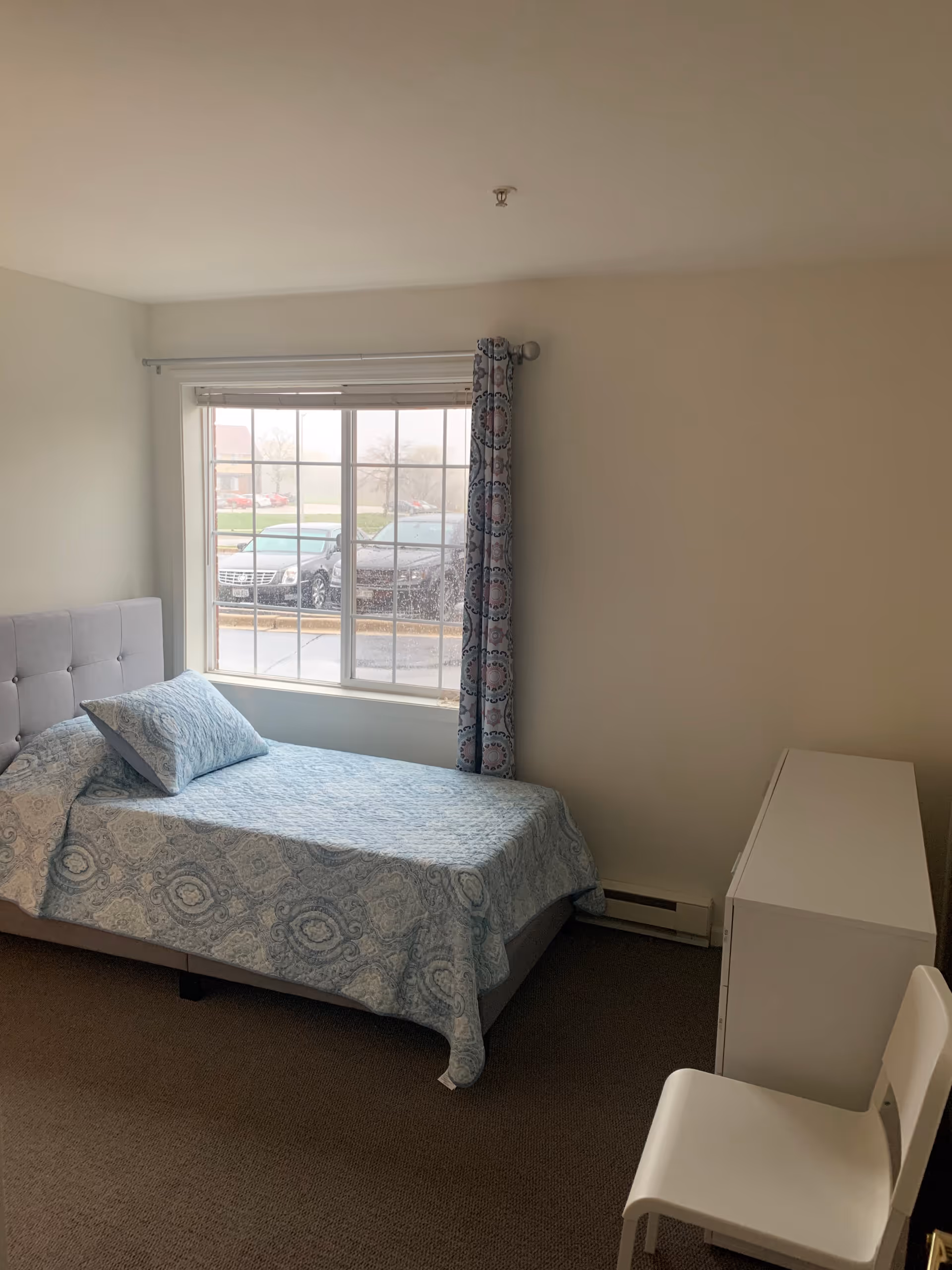 A simple bedroom with a single bed covered in a blue patterned bedspread and matching pillow. There is a large window with a curtain on one side, a white dresser, and a white chair. Outside the window, parked cars and some greenery are visible.