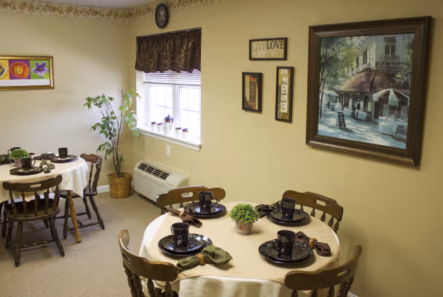 A cozy dining area with two round tables covered with beige tablecloths, each set with black plates, cups, and napkins. Four wooden chairs surround each table. The room has beige walls decorated with framed artwork and a small plant on each table. A window with brown curtains lets in natural light, and a potted plant is placed near the window above a wall-mounted air conditioning unit.
