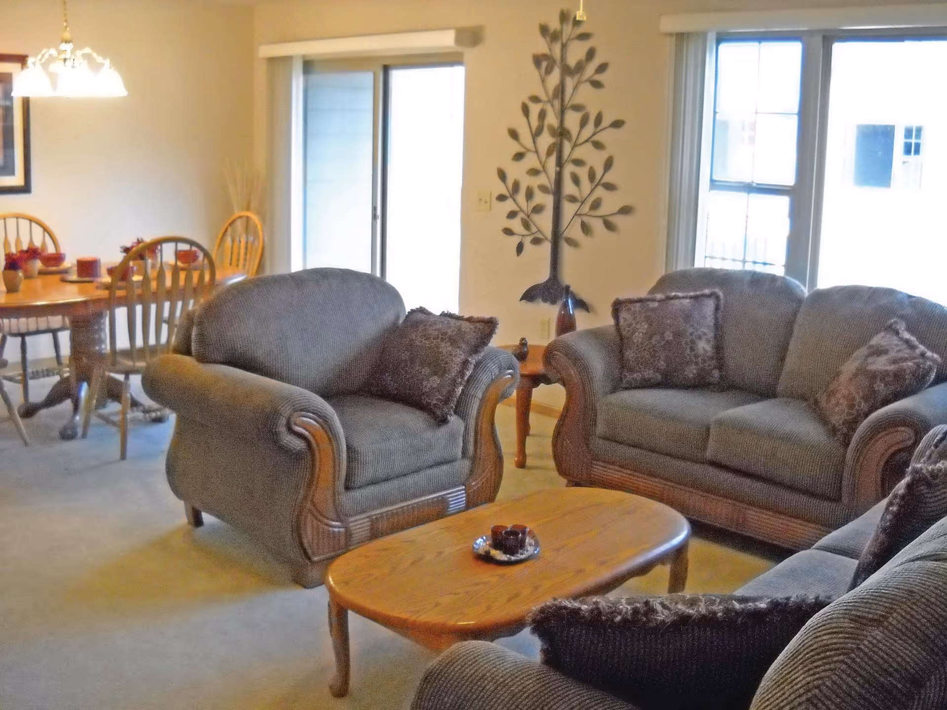 A cozy living room area with a matching set of cushioned armchairs and a sofa in a brownish fabric. There is a wooden coffee table in the center with a small decorative item on it. In the background, a dining area with a wooden table and chairs is visible, along with a sliding glass door and a window letting in natural light. A decorative metal tree wall art is mounted on the wall between the window and the sliding door.