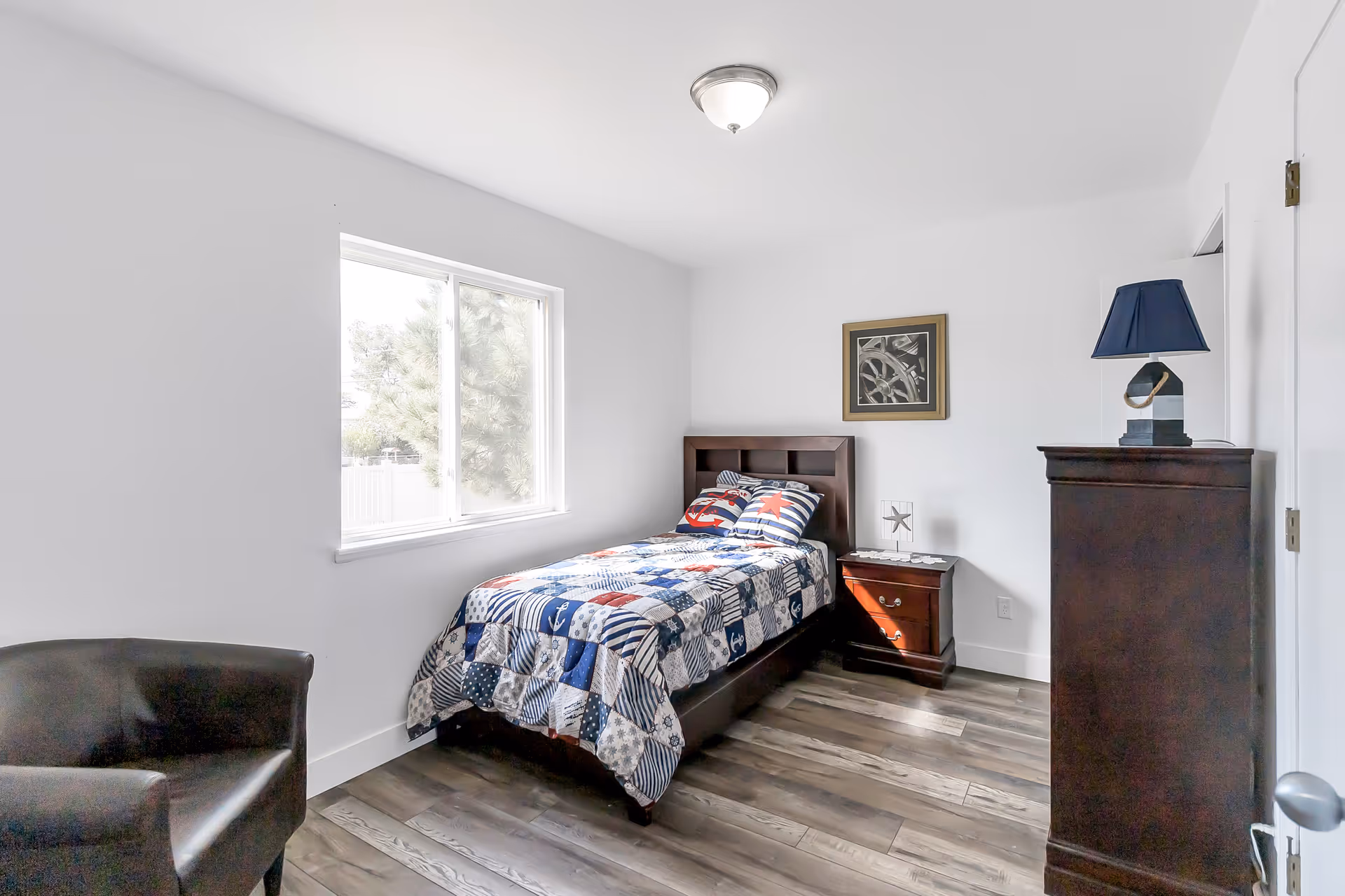 A tidy bedroom with a single bed, dresser, nightstand, and a window view.