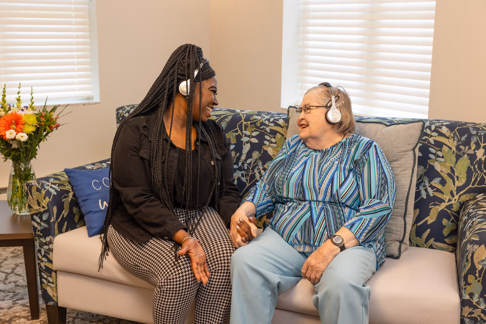 An elderly woman and a younger woman sitting on a floral-patterned couch in a well-lit room with window blinds. Both are wearing headphones and smiling at each other while holding hands. There is a side table with a vase of colorful flowers next to the couch.