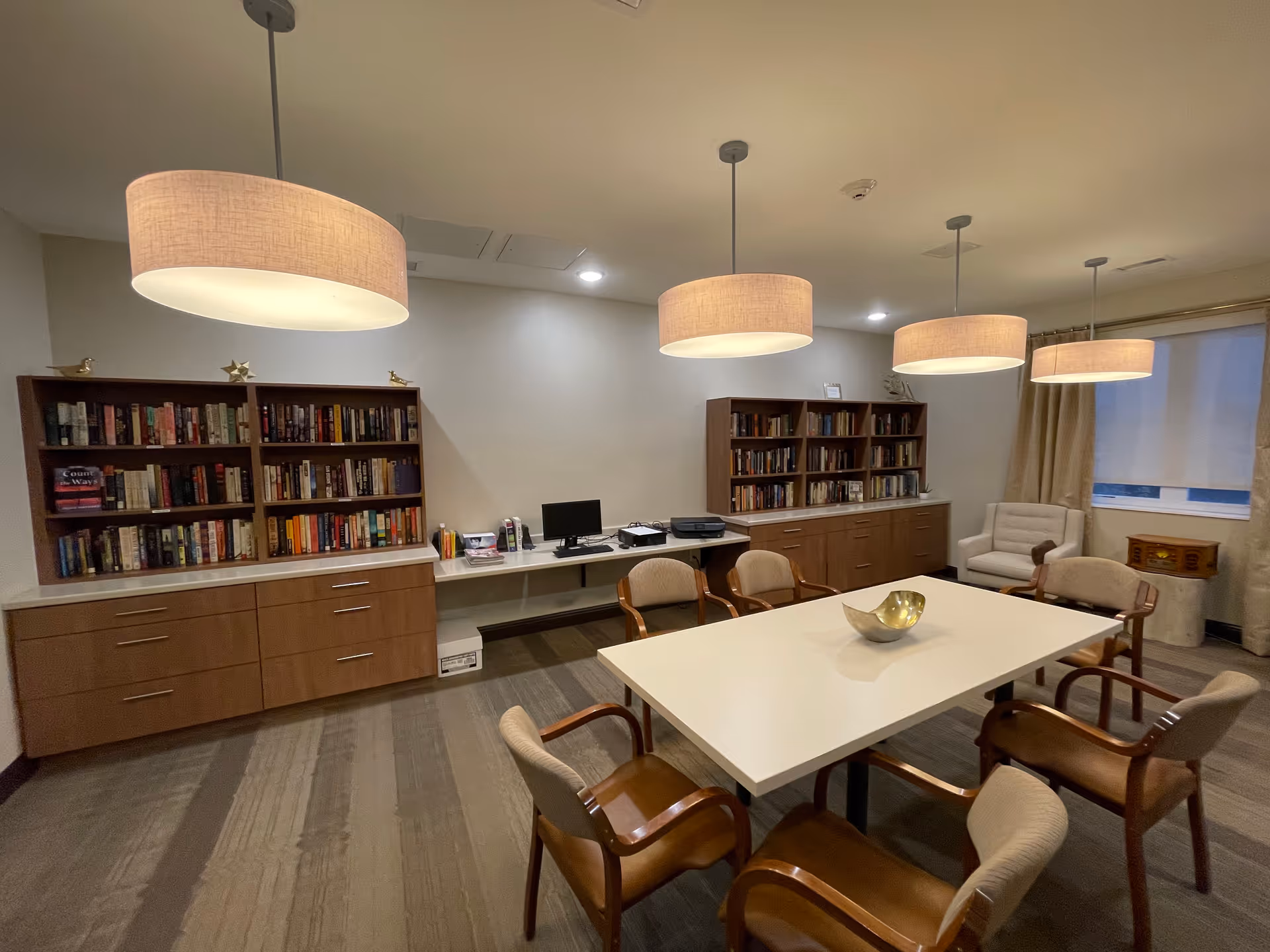 A well-lit room with a large white rectangular table surrounded by six cushioned wooden chairs. Two wooden bookshelves filled with books are against the wall, with a computer and printer on a desk between them. Four large pendant lights hang from the ceiling. There is a beige armchair near a window with curtains and a small wooden side table.