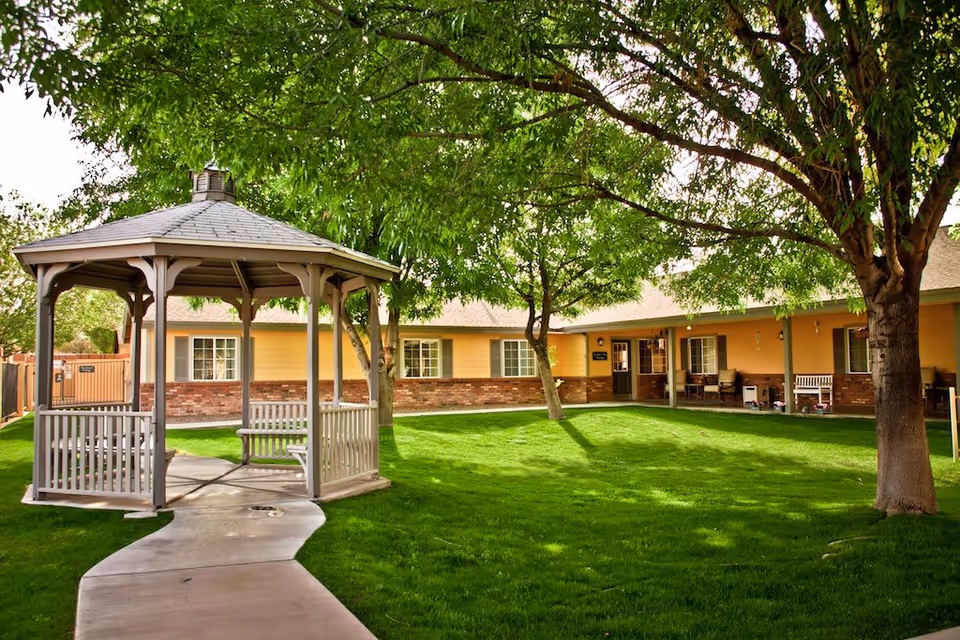 A peaceful outdoor courtyard at Cottages at Peoria featuring a wooden gazebo with benches, a concrete pathway, lush green grass, and several large trees providing shade. The background shows a single-story building with yellow walls, brick accents, and windows with white frames.
