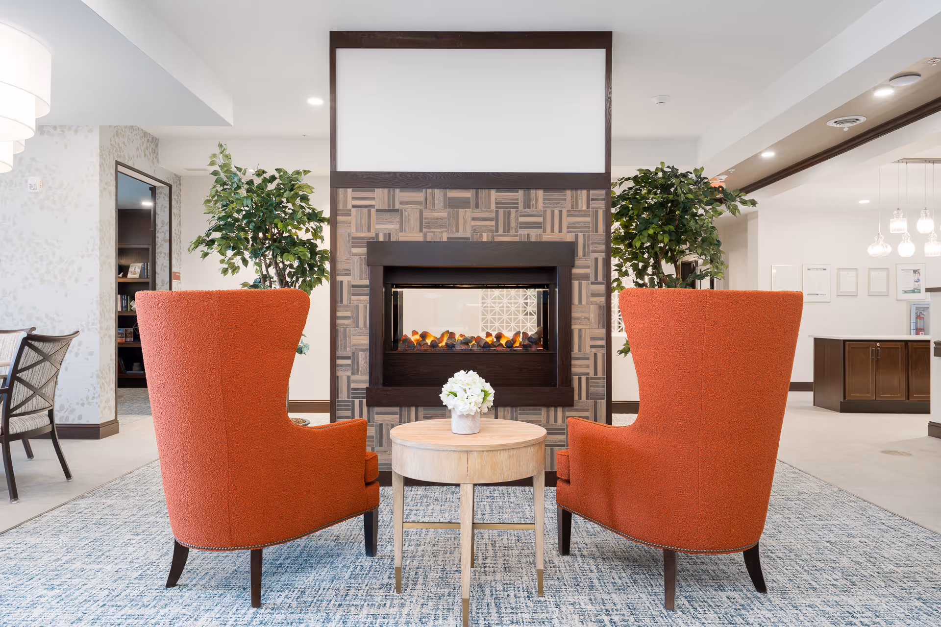 Two orange upholstered armchairs facing a modern electric fireplace with a wooden patterned surround in a bright, spacious room. A small round wooden table with a white flower arrangement is placed between the chairs. There are green potted plants on either side of the fireplace, and additional seating and lighting fixtures are visible in the background.