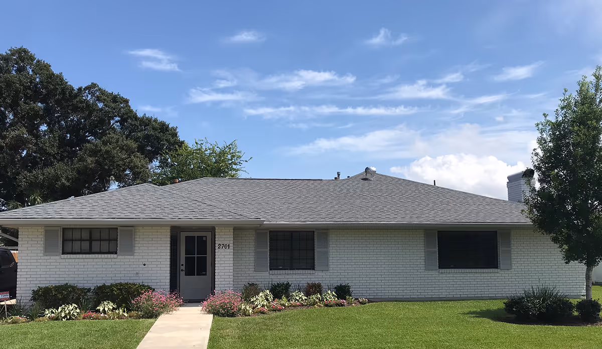 Single-story white brick building with a gray shingled roof, three windows with gray shutters, a central door, and a well-maintained lawn with flower beds and a tree on the right side under a partly cloudy blue sky.