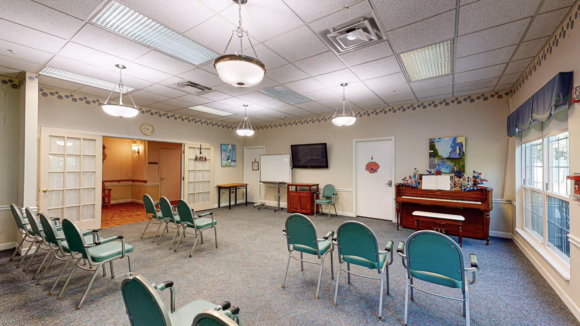 A community room with green chairs arranged in rows facing a piano and a wall-mounted TV. The room has carpeted flooring, white walls with a decorative border near the ceiling, and large windows on one side. There are three hanging light fixtures on the ceiling, a whiteboard, and a small table near the back wall. Double glass doors lead to another room.