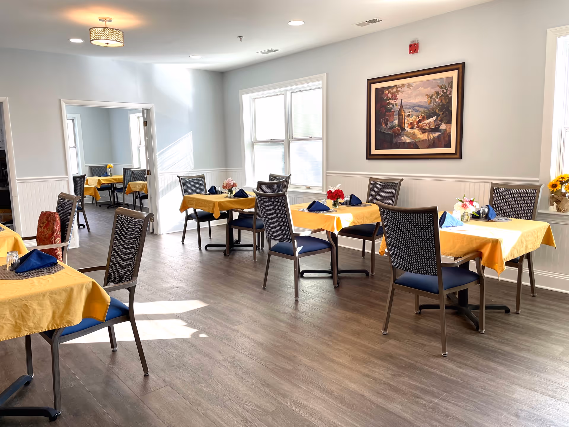 A bright dining room with several tables covered in yellow tablecloths and set with blue napkins. Each table has a small floral centerpiece. The room has light-colored walls, wood flooring, and a framed painting on the wall. There is a doorway leading to another room with similar tables and chairs.