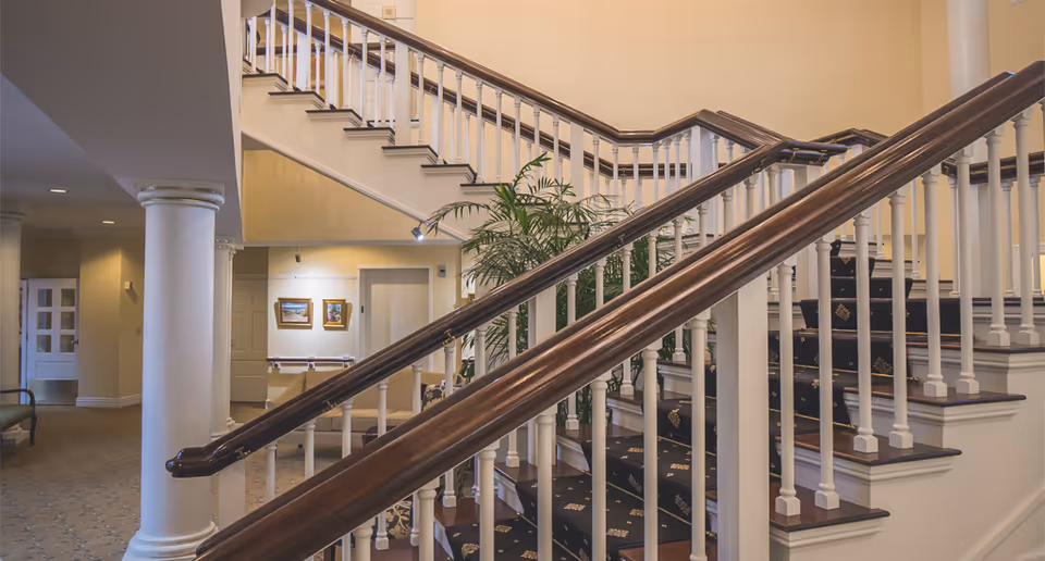 Interior view of a senior living facility showing a carpeted staircase with wooden handrails and white balusters. The area is well-lit with warm lighting, featuring beige walls, white columns, framed artwork on the walls, and a potted plant near the staircase.