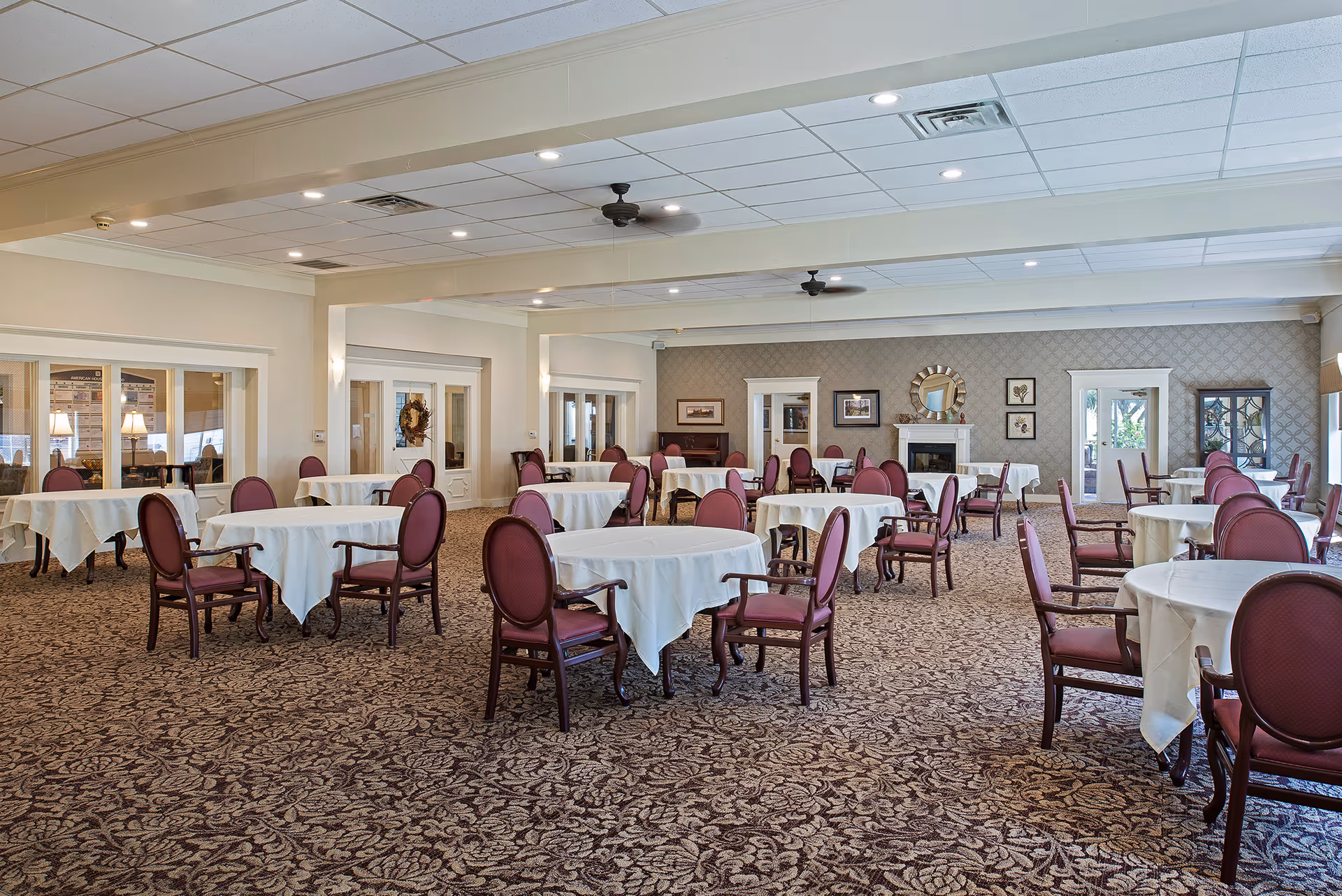 A spacious dining room with multiple round tables covered with white tablecloths, each surrounded by maroon cushioned chairs. The room features patterned carpet, a fireplace with a decorative mirror above it, framed artwork on the walls, and ceiling fans. Large windows and glass doors allow natural light into the room.