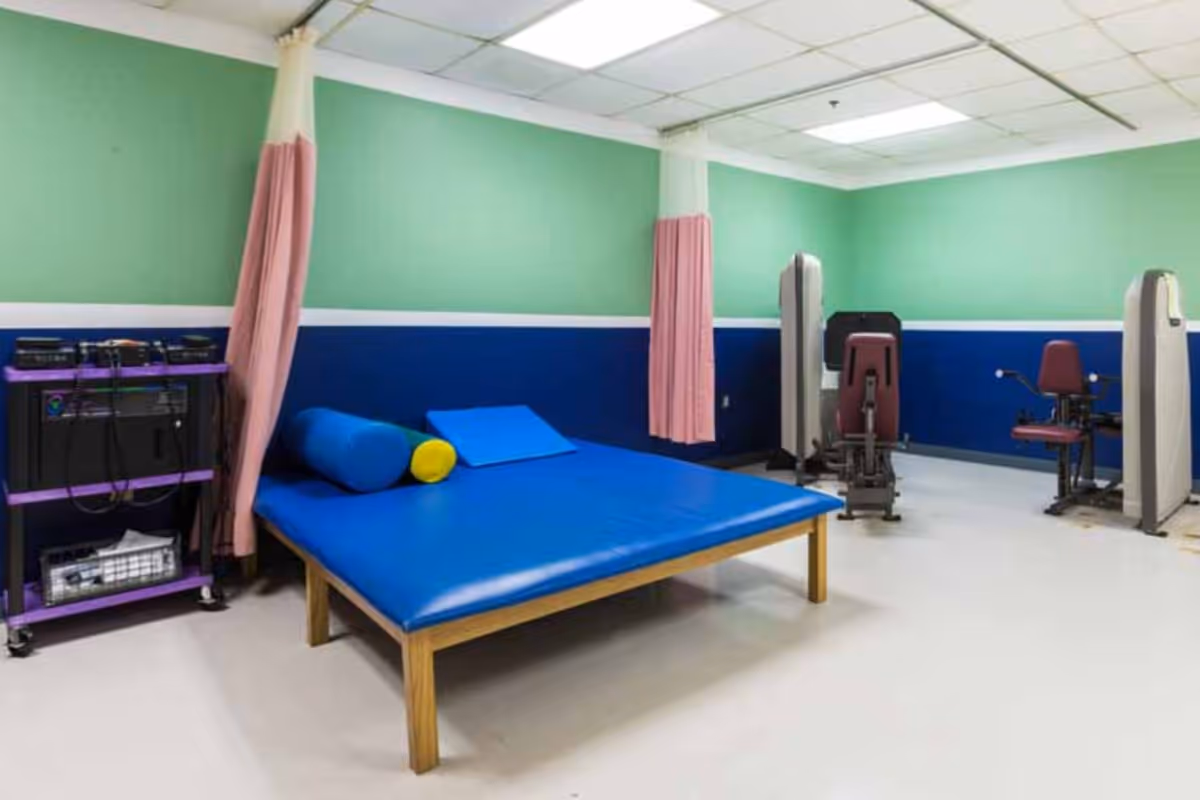 A physical therapy or rehabilitation room with a large blue padded table, cylindrical and wedge-shaped cushions, exercise machines, and a cart with electronic equipment. The walls are painted green and blue with a white stripe, and there are pink privacy curtains hanging from the ceiling.