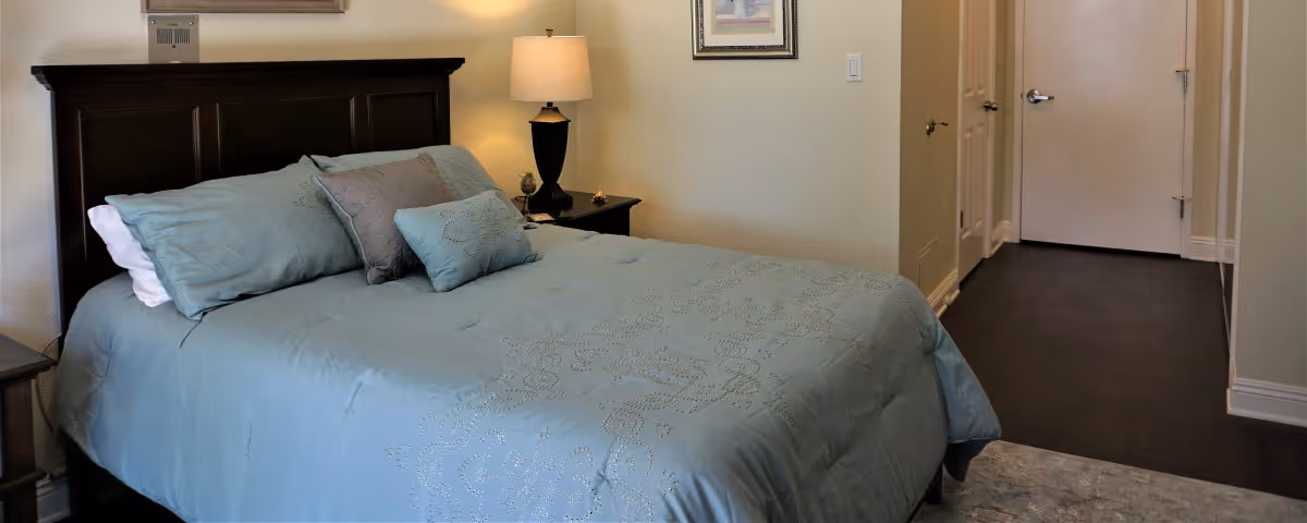 A tidy bedroom with a bed in light blue bedding, a bedside table and lamp, and a doorway opening onto a dark-floored hallway.