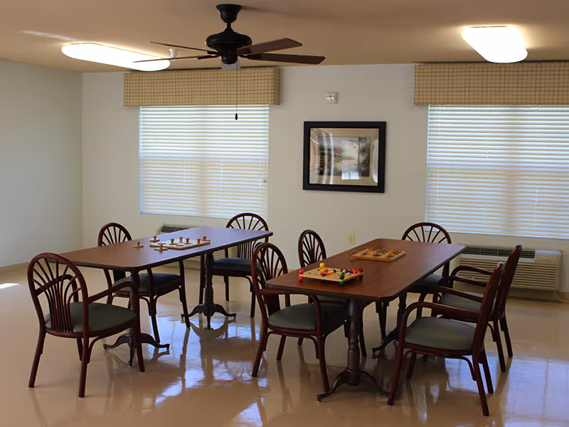 A well-lit room with two wooden tables and several wooden chairs arranged around them. Each table has a board game set up on top. The room has two windows with blinds and valances, a ceiling fan, and a framed picture on the wall.