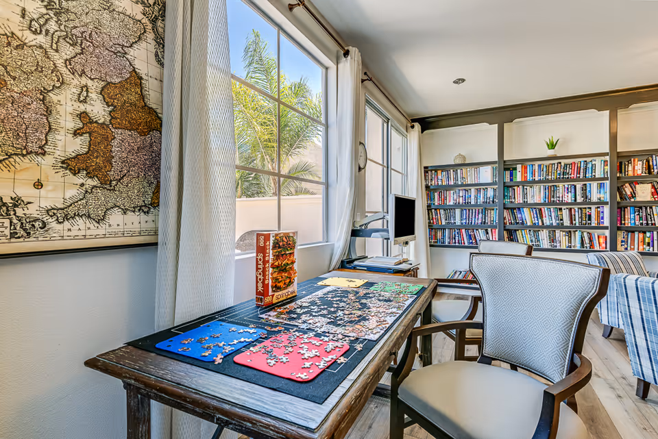 Sunny indoor activity room with a large bookshelf, puzzle table, and computer.