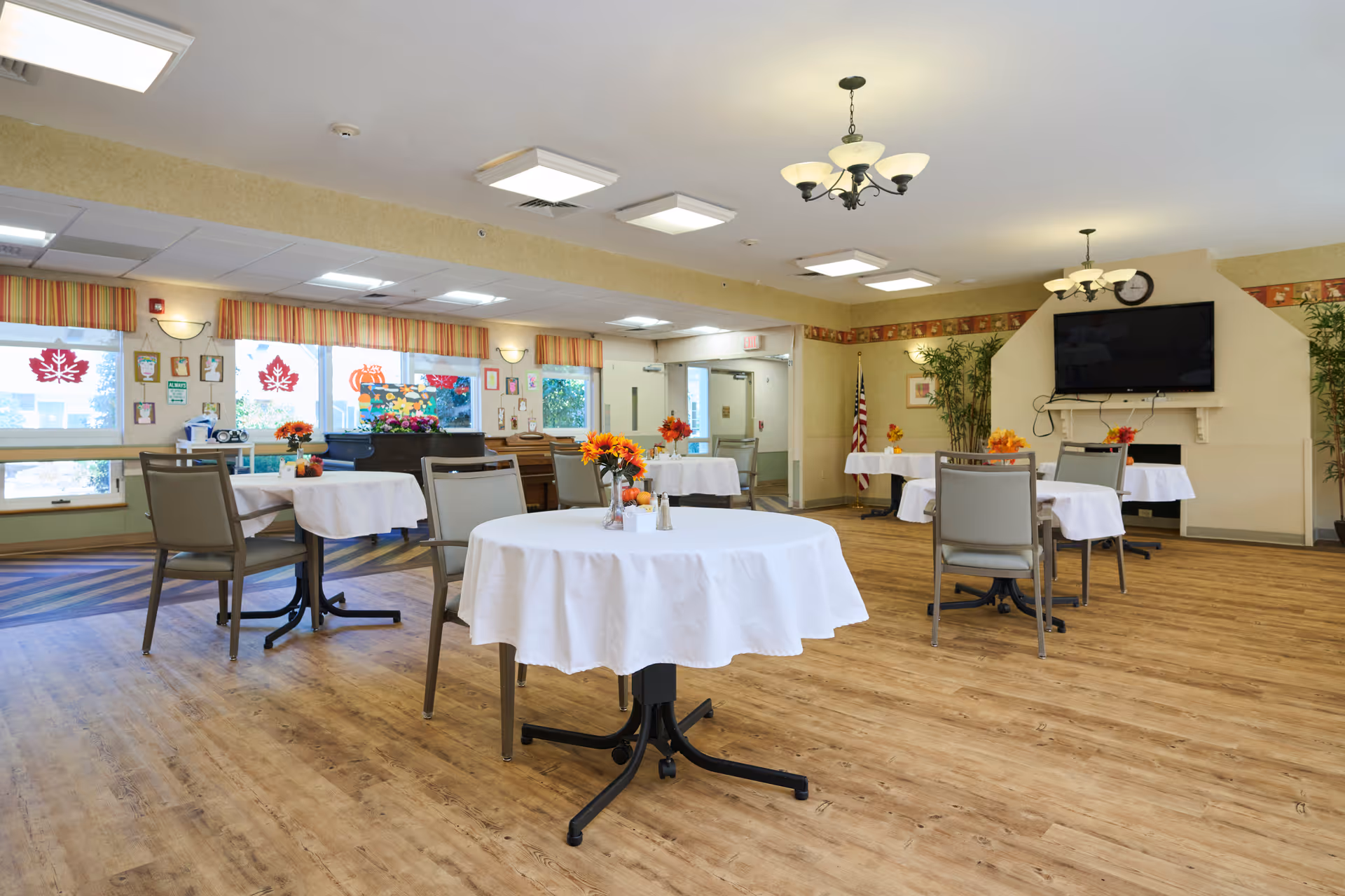 A bright and spacious dining room with several round tables covered with white tablecloths, each adorned with a small vase of orange flowers. The room has wood flooring, large windows with striped valances, a wall-mounted flat-screen TV, and ceiling lights including chandeliers. There are chairs around the tables and some plants in the corners, creating a welcoming atmosphere.