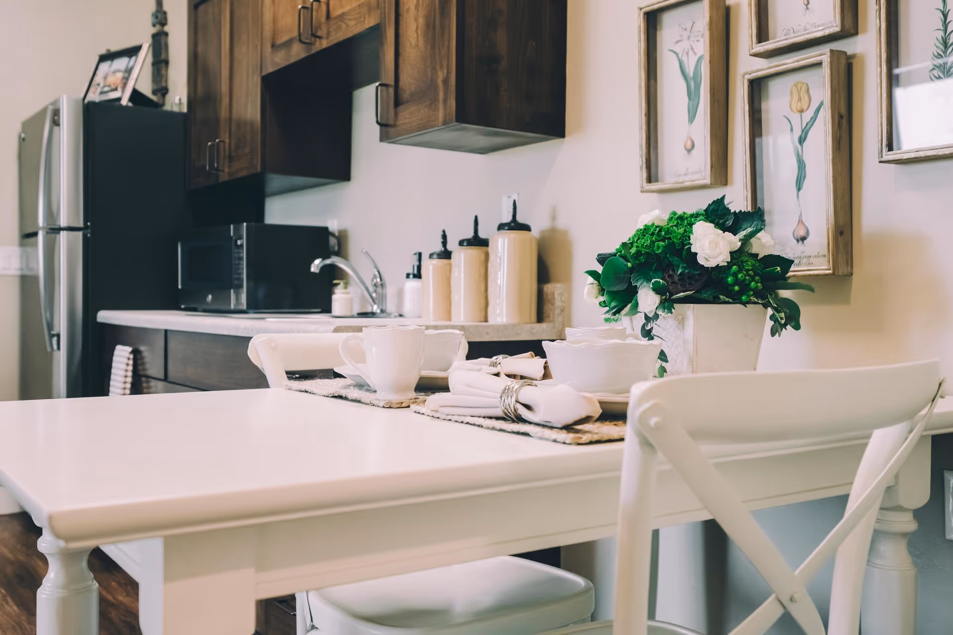 A kitchen area with a white dining table set with white cups, bowls, and napkins. The background shows wooden cabinets, a stainless steel refrigerator, a microwave, and decorative framed botanical prints on the wall. A green and white floral arrangement is placed on the table.