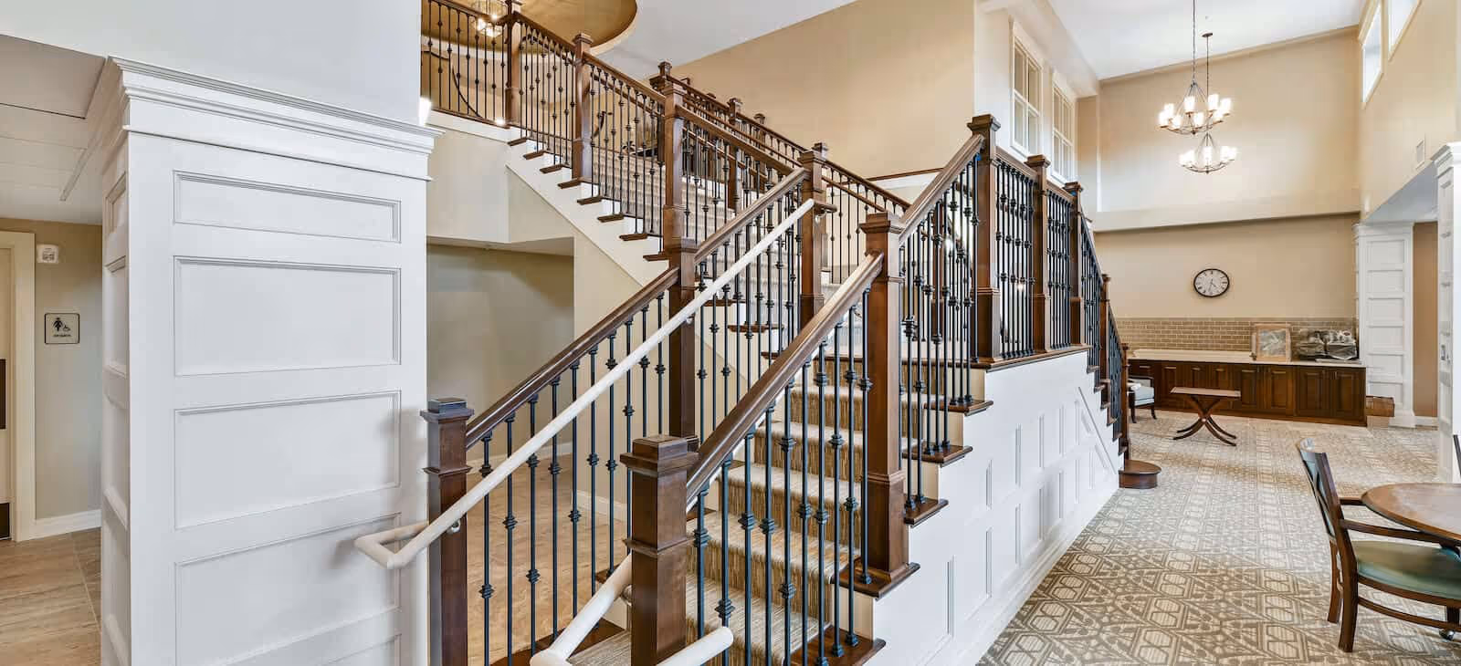 Interior view of a senior living facility featuring a wide staircase with wooden handrails and black iron balusters. The area has high ceilings with two chandeliers, patterned tile flooring, a round wooden table with chairs, and a wall clock above a cabinet in the background.