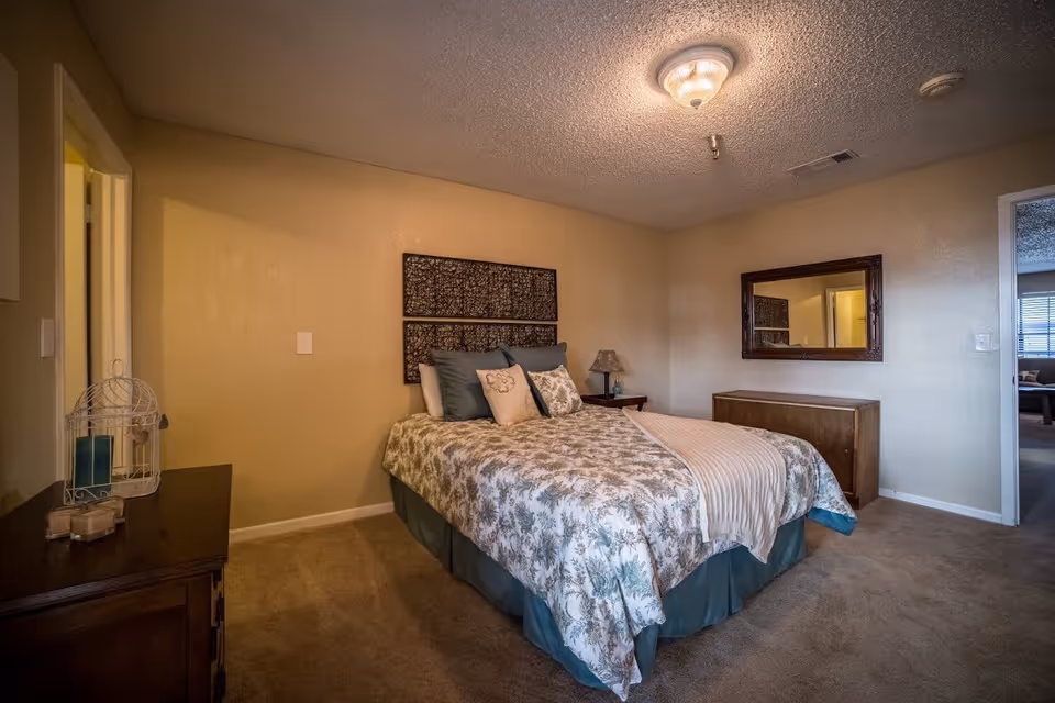 A cozy bedroom with a bed covered in floral bedding and multiple pillows. There is a wooden nightstand with a lamp and a small decorative item on the left side of the bed. A large mirror hangs on the wall above a wooden dresser. The room has beige walls and carpeted flooring, with a ceiling light fixture.
