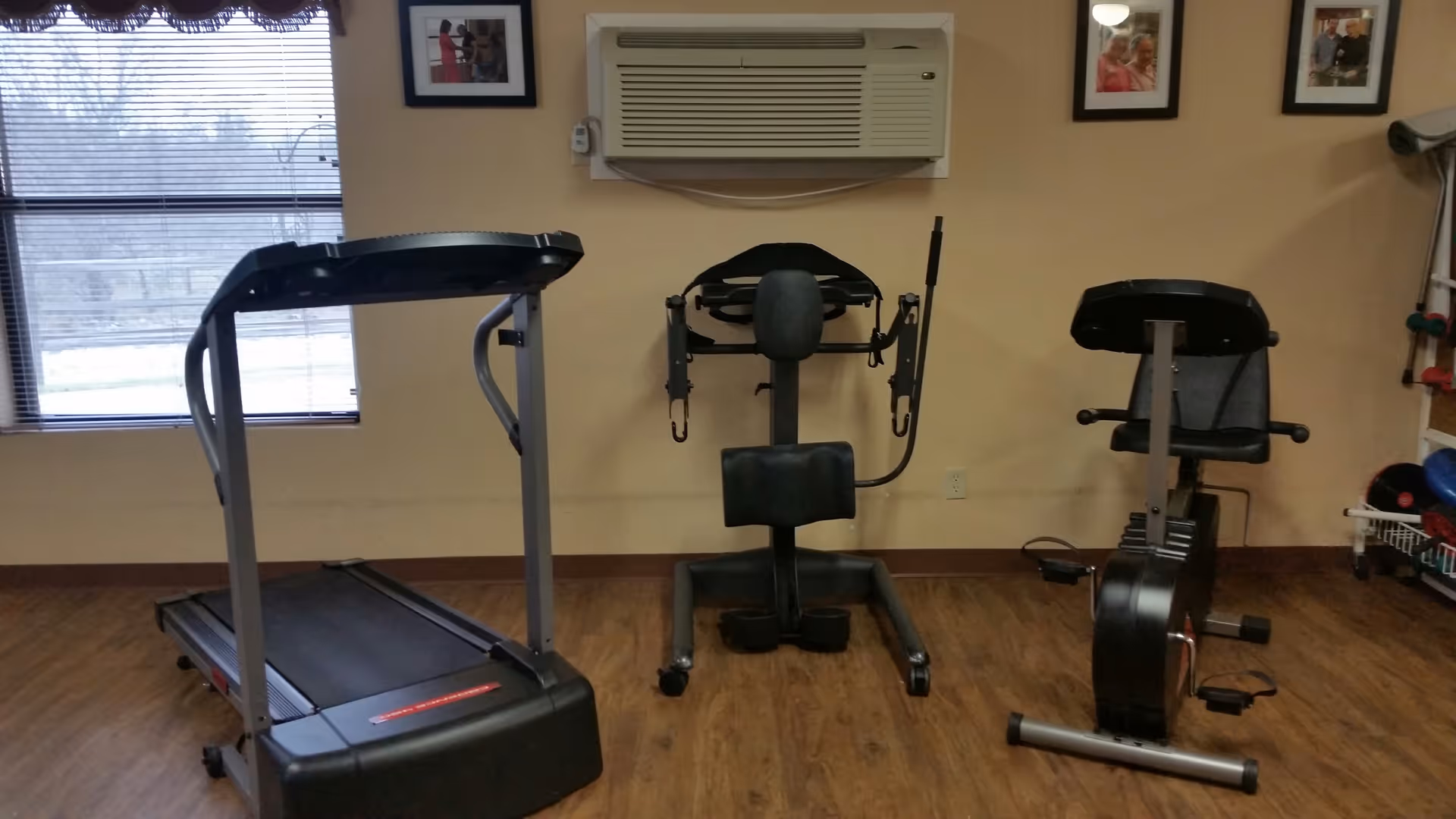 A small exercise room with a treadmill, a seated chest press machine, and a stationary recumbent bike. The room has wood flooring, beige walls, a window with blinds, and framed pictures on the wall. An air conditioning unit is mounted above the exercise machines.