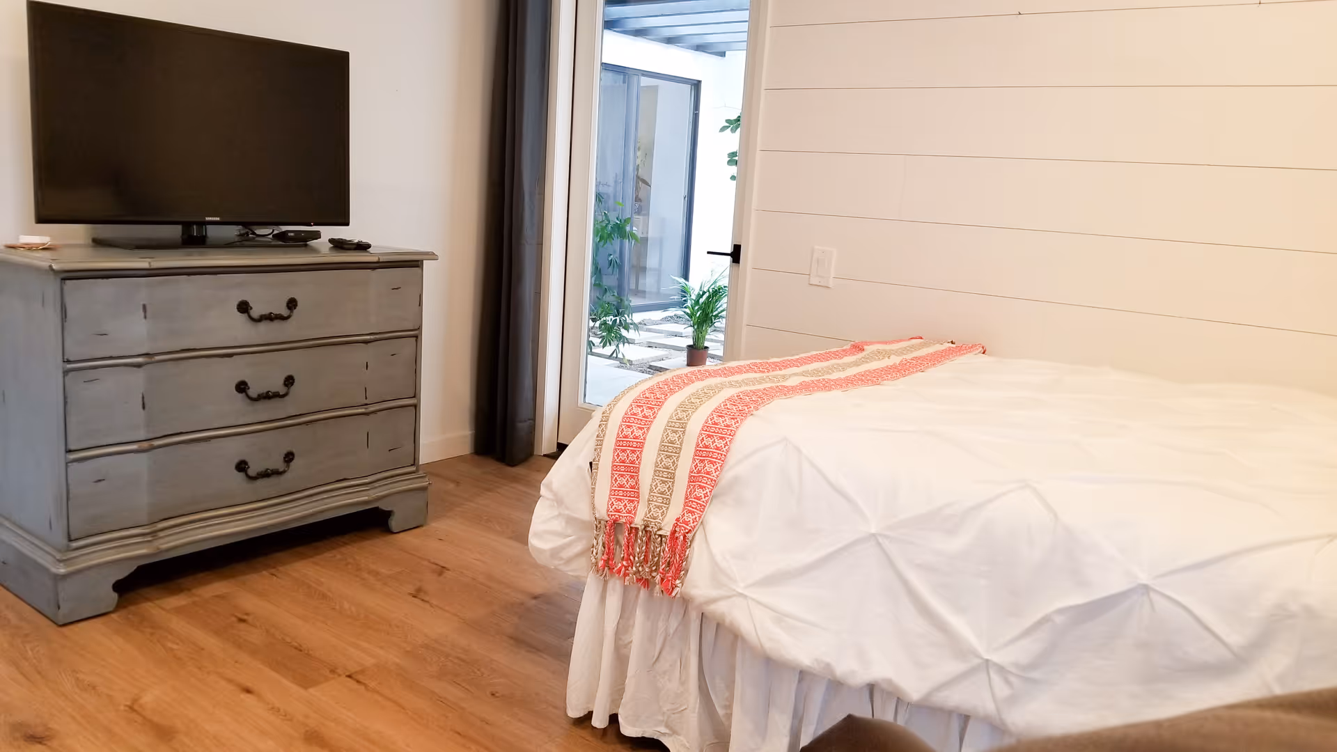 Bedroom with a white bed and red patterned throw, a gray three-drawer dresser topped by a TV, and a glass door opening to a small outdoor area.