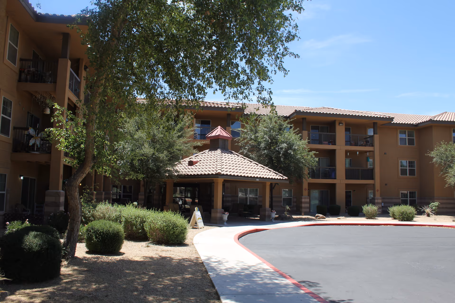 Exterior view of a senior living facility with a three-story building featuring balconies and windows. In front, there is a covered entrance with a tiled roof, surrounded by trees and bushes under a clear blue sky.