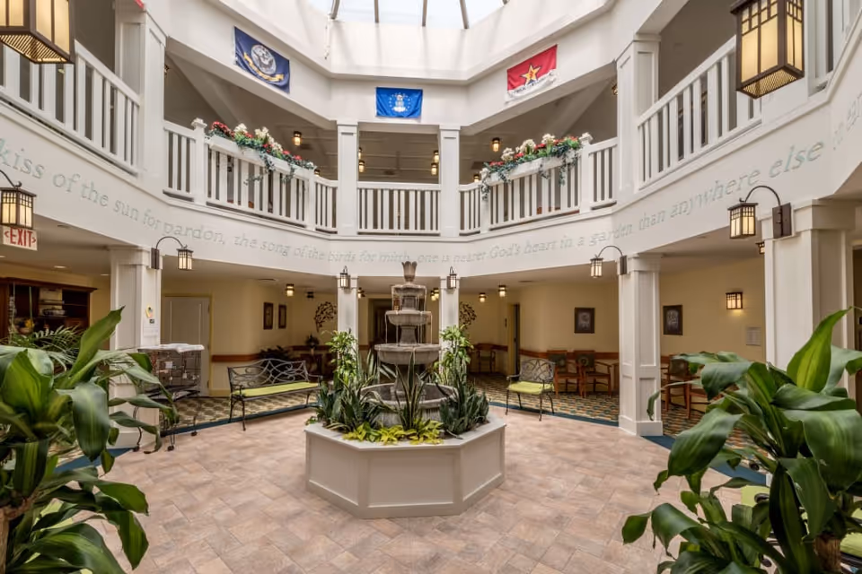 Interior view of a senior living facility atrium with a central water fountain surrounded by plants and benches. The space features a two-story design with white railings on the upper level, hanging lantern-style lights, and flags displayed above. There are seating areas with chairs and tables along the walls, and a skylight above provides natural light.