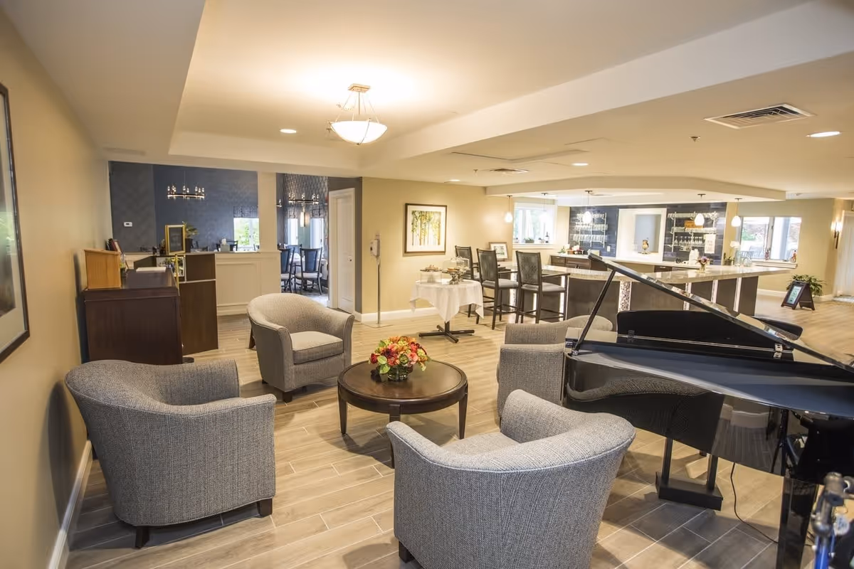 A cozy common area in a senior living facility featuring four gray upholstered armchairs arranged around a round wooden coffee table with a floral centerpiece. To the right, there is a black grand piano. In the background, there is a bar area with high chairs and pendant lighting, as well as a dining area with tables and chairs. The room has warm lighting, wood-look tile flooring, and neutral-colored walls with framed artwork.