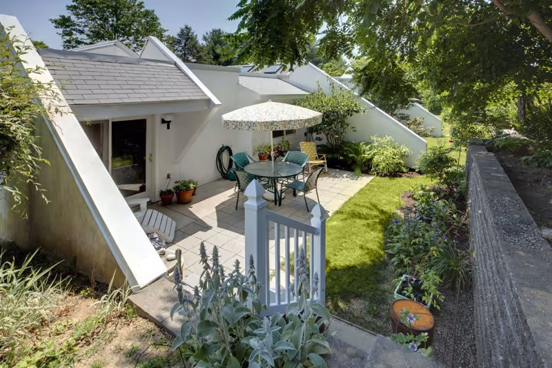Private ground-level patio with a round table and umbrella, chairs, potted plants, and a small lawn beside a white angular building.